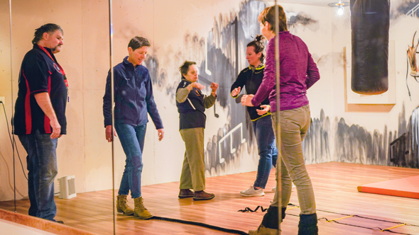 Mehrere Personen in einem Trainingsraum, zwei davon in der Mitte machen einen Balanceübung und laufen über ein am Boden liegendes Seil